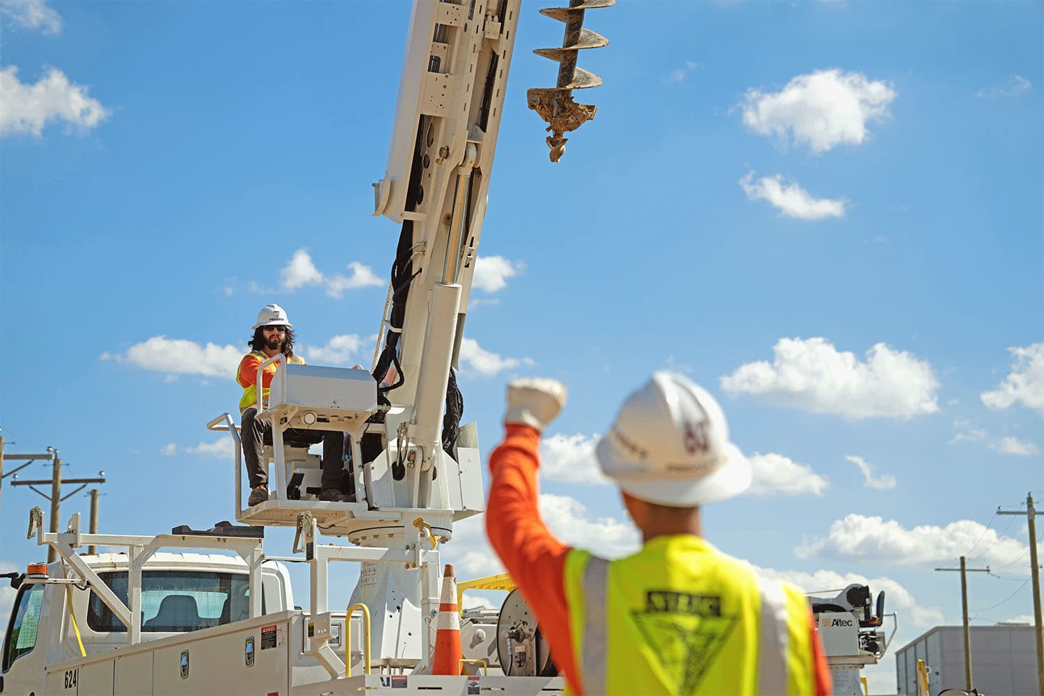 Student uses hand signal to another student operating a digger derrick